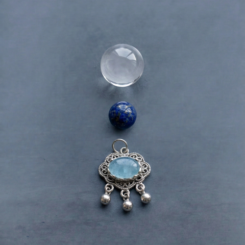 AETHER Key: CLARITY clear quartz bracelet deconstructed detail shot, featuring one flawless transparent clear quartz bead, one deep lapis lazuli bead with pyrite flecks, and one intricate silver cloud-pattern pendant with a faceted light blue crystal, macro perspective on a cool grey background.