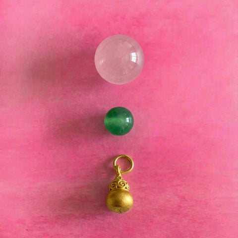 AETHER Abundance Collection - Vibrant minimalist vertical arrangement featuring a pale pink crystal sphere, a vivid green gemstone bead, and an ornate oriental gold gourd pendant, set against a bright pink background.