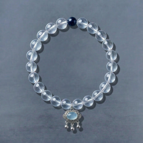 AETHER Clarity Collection - Natural clear quartz crystal bracelet featuring a dark blue lapis lazuli bead and an ornate silver pendant with aquamarine, styled on a minimalist dark grey background.