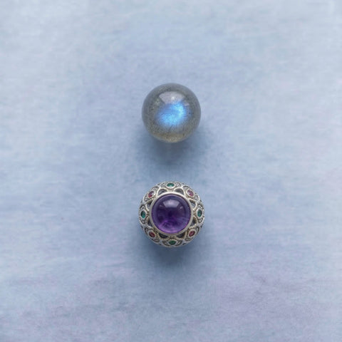 AETHER Jewelry Details - A minimalist vertical close-up of a natural labradorite bead with strong blue flash, placed above an intricate silver focal bead with a purple amethyst center, on a soft pastel blue background.